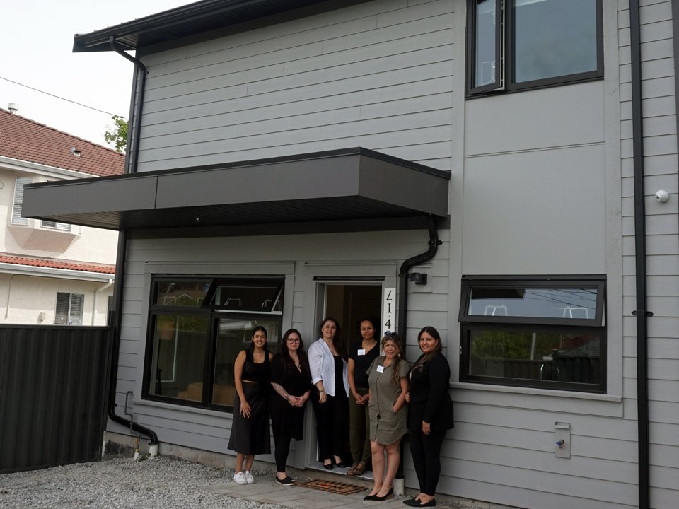 Four women from Aunt Leah's Thresholds staff pose standing outside a two storey house. The women are smiling and dressed in black and other neutral colors. The house has three windows and the door is open.